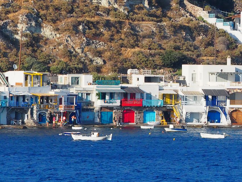 A row off houses, all connected to each other, 2-3 stories tall. The ground floors all have a brightly-painted large door - once used to store boats. UPstairs they all have a balcony painted in the same bright color, and doors and window have the same color. Other than the colored parts, the houses are all white. A few boats bob at anchor in front of the houses.