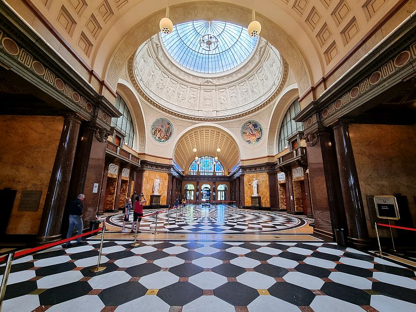 A long hallway, straight sided except the middle portion, which is round, with rounded sides and a large dome above it. Part of the dome is visible in the view and shows decorative detail around the bottom of the dome and the top of the dome is panes of glass. The floor is marble placed in a chessboard pattern. The walls sport pillars of marble at each doorway. Many historic spa towns had such ornate gathering places.