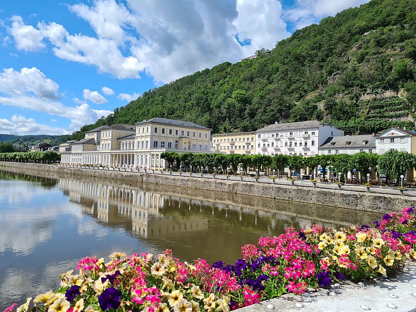 Looking along the river at the right side of the river: nearest is a plot of very neat trees, with a row of buildings visible behind them. Beyond the trees, edging the river, is the Kuursaal. Quite a large building, three stories tall but extending quite far along the bank. Arched windows on the ground floor with pillars between them. Painted a light yellow with white trim. Behind the row of buildings rises a green hill. 