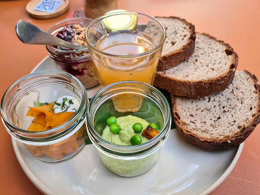 Three slices of round brown bread on the right, arranged to overlap. In the front, pea mush with a few whole peas on top in a small jar without a top. Left of that, a salad of some sort, mostly carrot, also in a small pot. Beyond that on the left, oatmeal with some berries on top and a spoon sticking out, also in a small pot without a lid. A glass of apple juice in the center. A jar of jam is behind, but only the lid is visible.