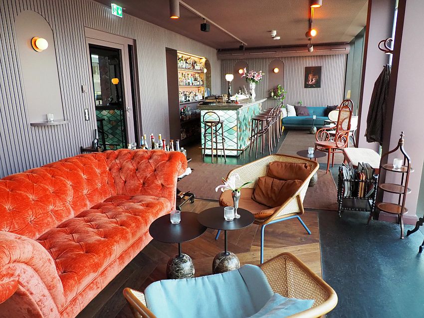 In the background is the bar with bar stools lined up along it. In the foreground, an orang sofa that appears to be upholstered in velvet and two ratan chairs opposite it with cushions. Other such chairs are visible along the wall and another sofa, blue this time, beyond the bar in the background. Coffee tables, a magazine rack, a coat tree, etc. make it look very homey.