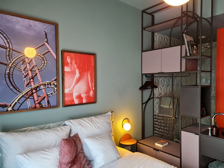 This view shows the top end of the bed with several pillows against the backboard. The wall above the bed has two framed photos: one shows part of a metal roller coaster track. The other shows a woman from the back looking in the mirror, wearing a tight tee-shirt and no underpants. A wall unit along the side of the bed has open shelves and a couple closed cupboards, as well as a rod and some hangars. Part of a sink is also visible in the wall unit. Behind the wall unit (as seen through the wall unit), the wall is patterned tile, white but with a profile. 