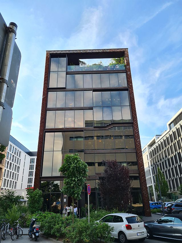 A rectangular building, not very wide, but about seven stories high. The front is glass and reflects the building opposite, that also looks like an office building. Around the stories and the sides of the building are in dark red brick or stone. The top floor has a balcony of sorts that does not have a roof, so it is a partial roof garden, even if it's not on the roof. On the street, a few trees and bushes, parked cars and bicycles.