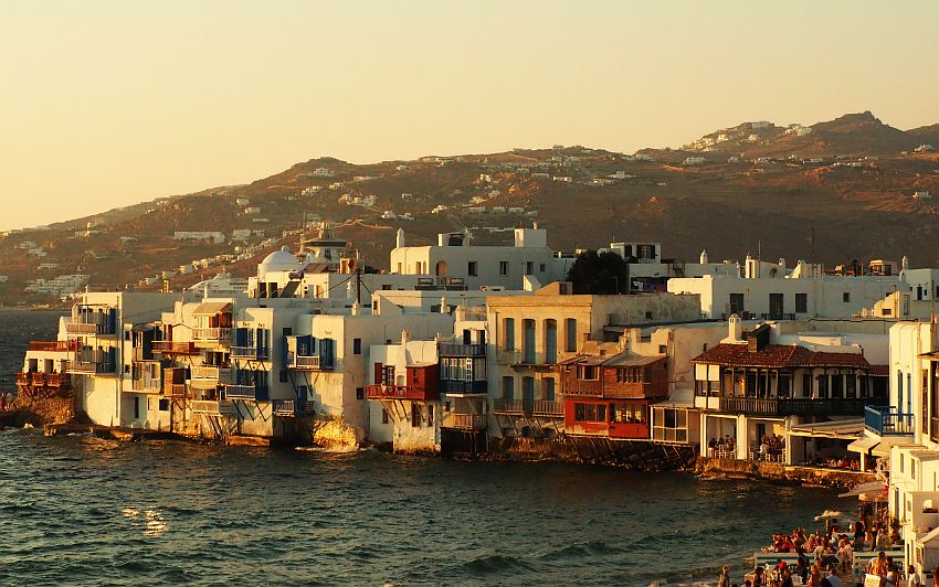 A row of buildings, mostly about 3 stories tall, mostly white, along the water, i.e. there is no beach, just the water lapping at the foundations of the buildings. Many have balconies, and some are clearly restaurants because people are visible sitting at tables in the openings. Bottom right of the picture more restaurants or cafes, crowded with people. Beyond the buildings are some hills, dotted with white houses.