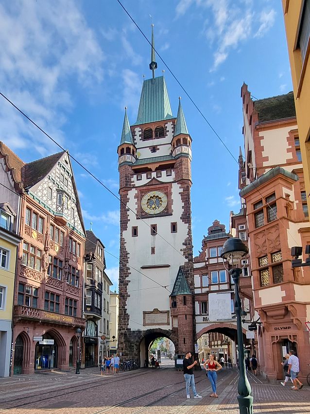 The tower has an archway through it at the bottom, and the smaller building next to it also has a large archway under it. The tower is white ut has brick at the corners. A clock toward the top, a turret on each corner and a steep green roof on top.