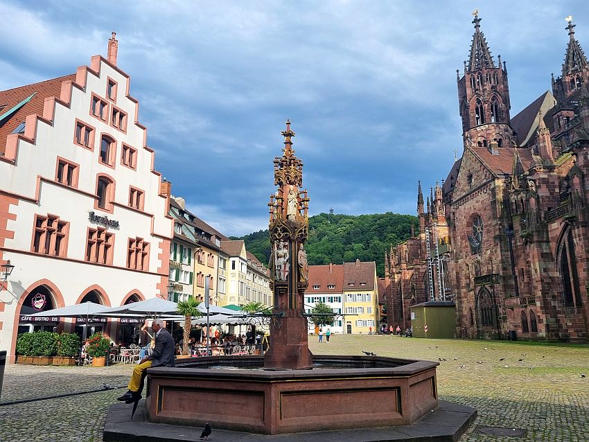 In the center, an ornate fountain with a vertical pole in the center, and walls around the bottom to hold the water. Left, a row of buildings in light pastel colors and outdoor cafes in front. Ahead, the plaza is mostly empty. On the right, the gothic and Romanesque side of the cathedral.