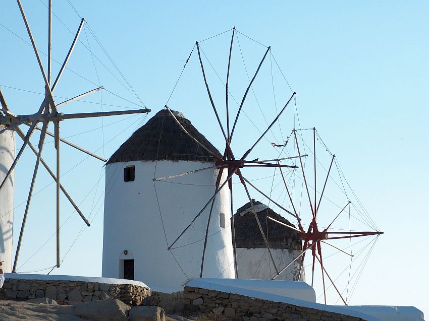 3 white, cylindrical windmills with pointed straw roofs. what used to be the windmill part is just an array of wooden poles in a circle.