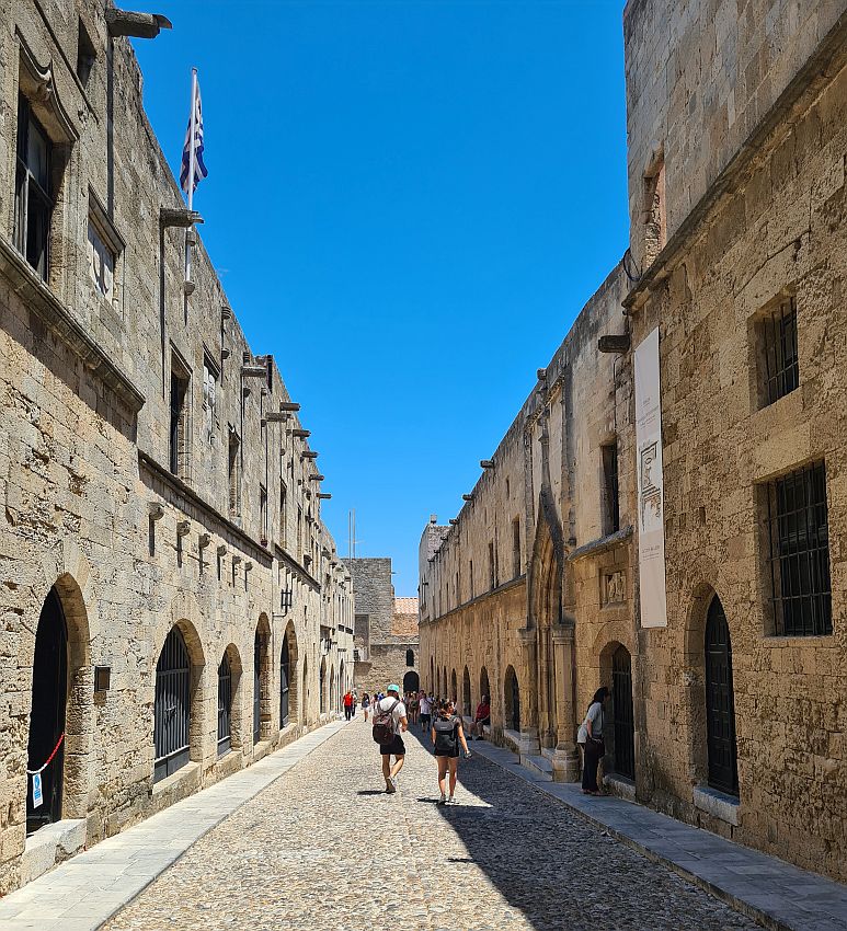 Old stone buldings on either side of a cobbled street. The buildings are mostly 2-3 stories tall and have arched entryways on their ground floor: mostly rounded romanesque arches, but one large gothic arch is also visible on the right.