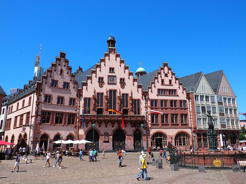 Each of the three buildings has a stepped gable up to a point. They're all painted pale pink while the trim is a darker pinkish-red. The ground floor entries and windows all have arches over them. The middle building has small sculptures between and around the windows on the upper stories. In front of them on the square, people pass by and there is a fountain with a statue of justice holding a scale.