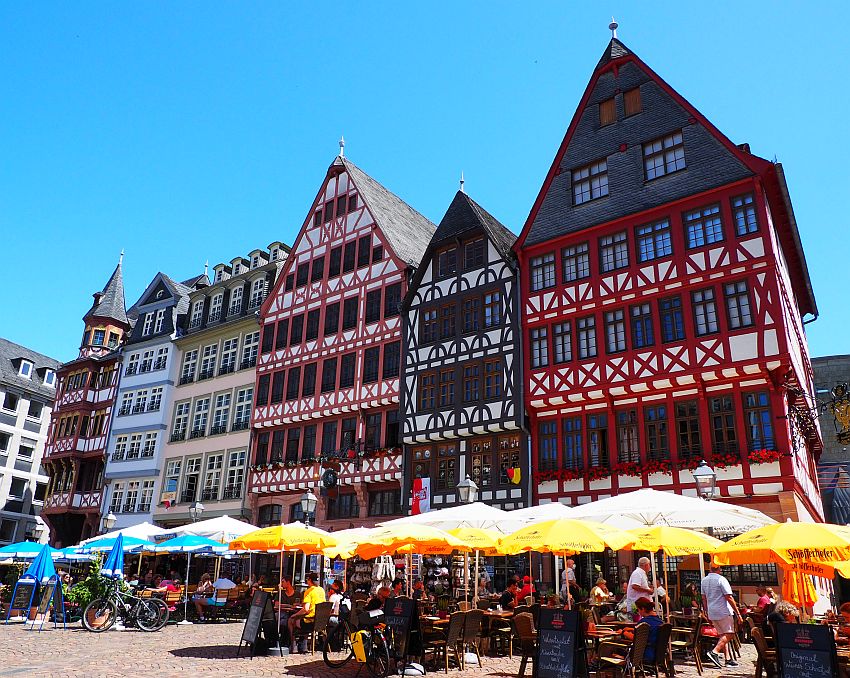 On the Romerberg a row of rebuilt medieval buildings. They're all 5-6 stories tall, with timbered facades. The timber is painted in colors while the spaces between the timbers are white. Most have peaked roofs. In front of them on the square is a row of outdoor cafes with large umbrellas shading the tables.