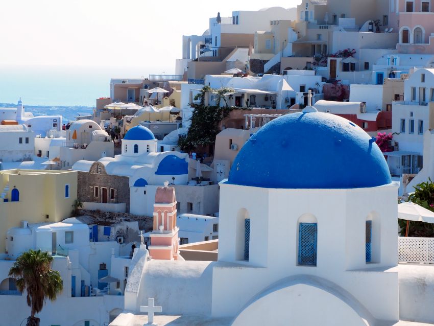 A view over a jumble of mostly white buildings, some with blue domes and blue doors. The nearest building is a blue-domed church.