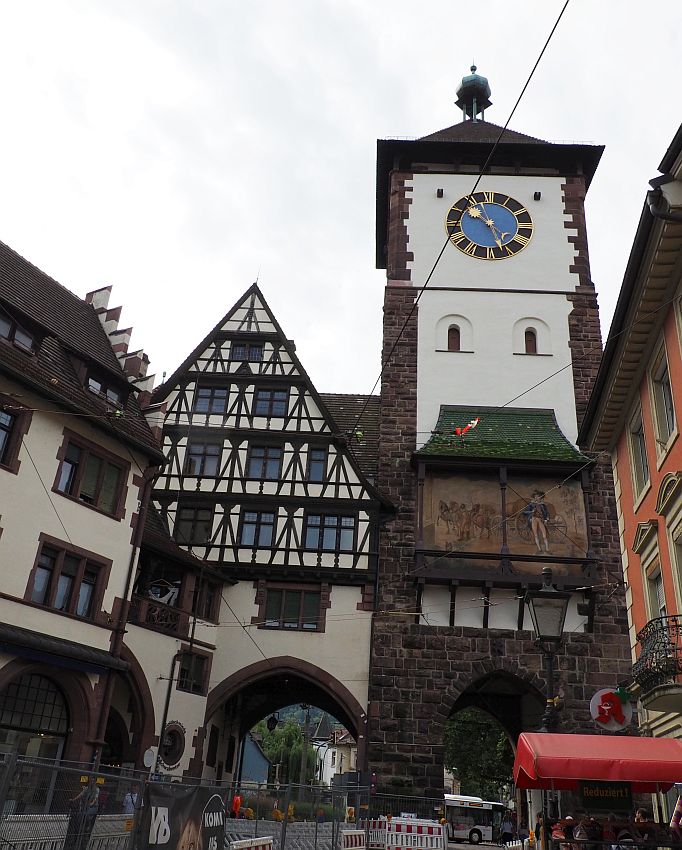 The tower is square and white, with brick on the corners. There's a big clock just under the rather plain roof. Lower down is a large painting of a person and some horses - the painting spans the width of the tower. Next to the tower is a half-timbered building with a pointed roof. Both buildings have large archways on the ground floor, big enough to let cars through.