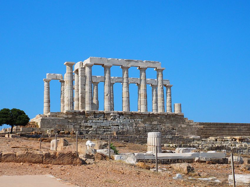 The temple stands on a high stone base against a blue sky. On the base is a square of lined-up columns, some of them holding up cross-stones as well. It is all white marble. Some additional pices lie on the ground in the foreground.