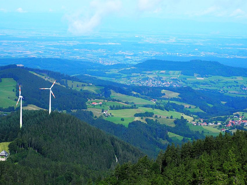 Foreground: the tops of tree-covered hillls, the left one has 2 large windmills on top. Beyond that, hilly terrain with a mix of green farmland, small villages and wooded areas. In the background, more of the same mix, but less distinct because of the distance.