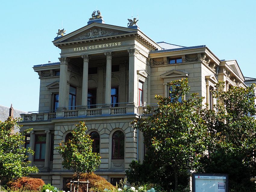 A brownish color stone siding. PIllars hoding up a pediment on the upper floor, with a bas-relief in the pediment and statues of winged lions above that on the corners of the pediment. The rest of the building is somewhat simpler.