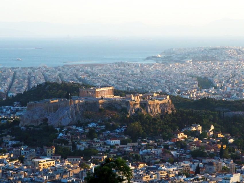 A view over Athens shows lots of white buildings, densely packed, on mostly flat ground, with the sea in the background (dim because of the haze). In the middle, a large hill, green around its edges but cliffs within that, and the acropolis and other ruins visible perched on the cliffs.