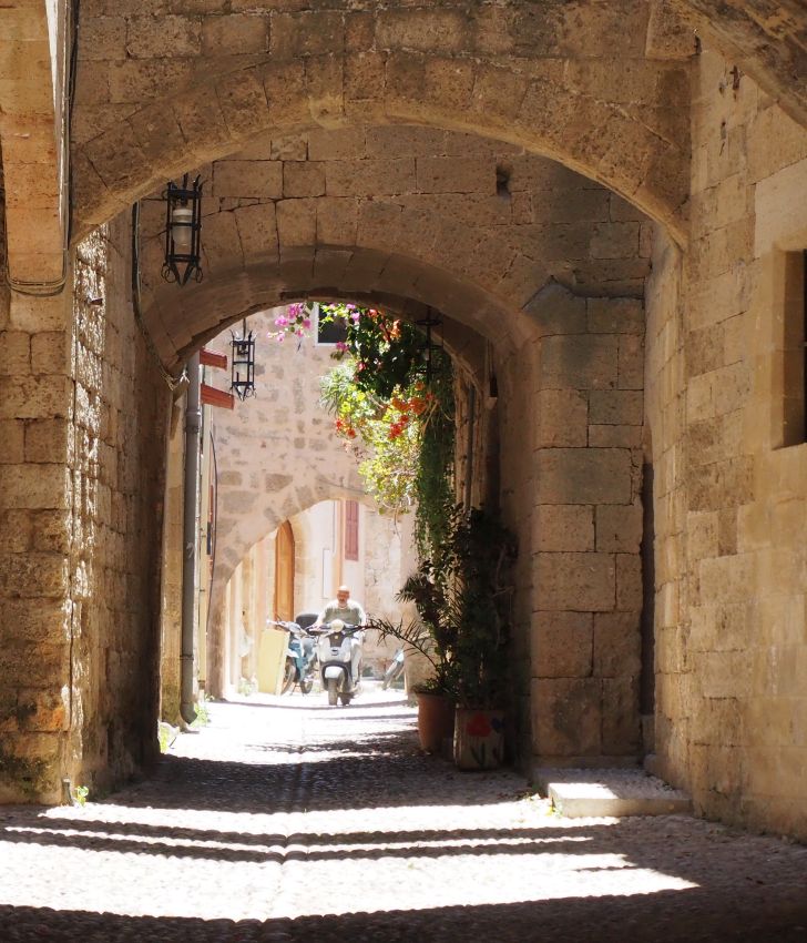 Looking along a narrow street: stone walls of buildings on both sides, arches over the street in the nearer part, then sun shining beyond that. The ground is cobbled. A motorcycle approaches.