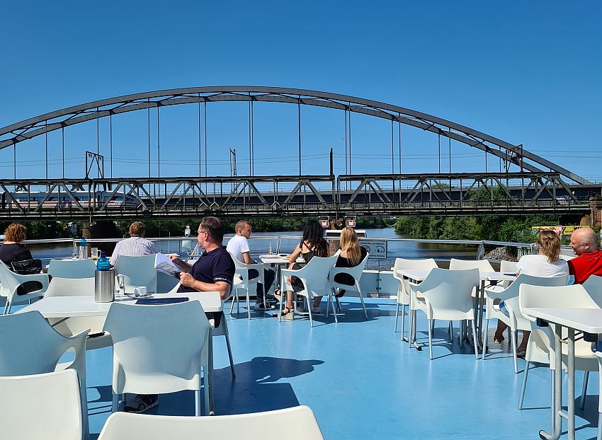 The deck of the boat is blue matching the color of the sky above. White tables with white chairs are set around the deck, and people sit at some of them. Across the photo in the middle, and seemingly at the same height as the deck, is an iron railway bridge. A train can be seen crossing it.