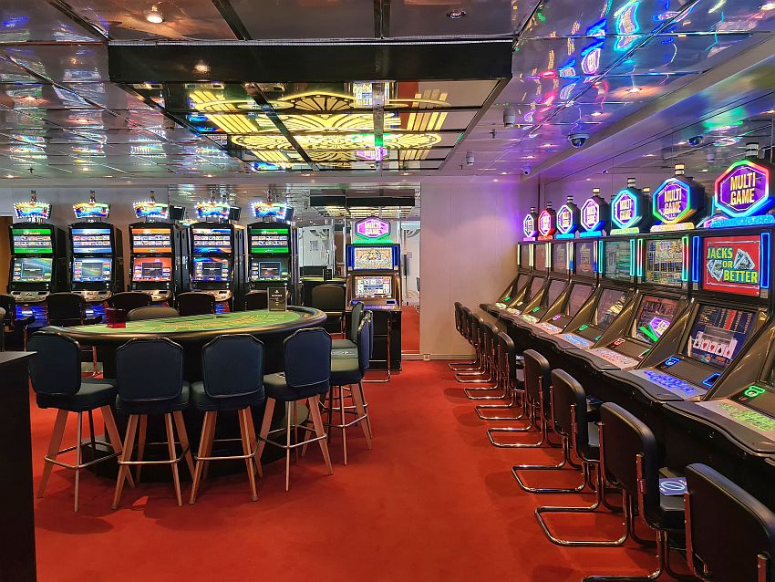 On the right wall and the back wall, rows of slot machines, each with a chair in front of it. In the middle of the room, semi-circular table with chairs around its curve. It looks like it's intended for blackjack. The slot machines all are lit up with bright lights, which reflect from the shiny ceiling.