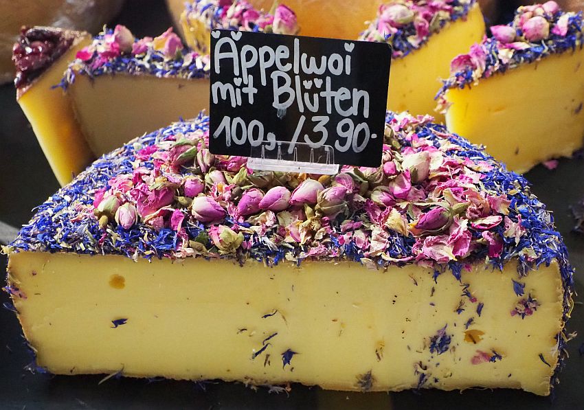 A half-round of cheese, cut so the cheese is visible, but the top is covered with what looks like rose buds and some other bluish-purple small flower. The sign on top of the cheese reads Appelwoi mit Bluten 100 g/3.90.