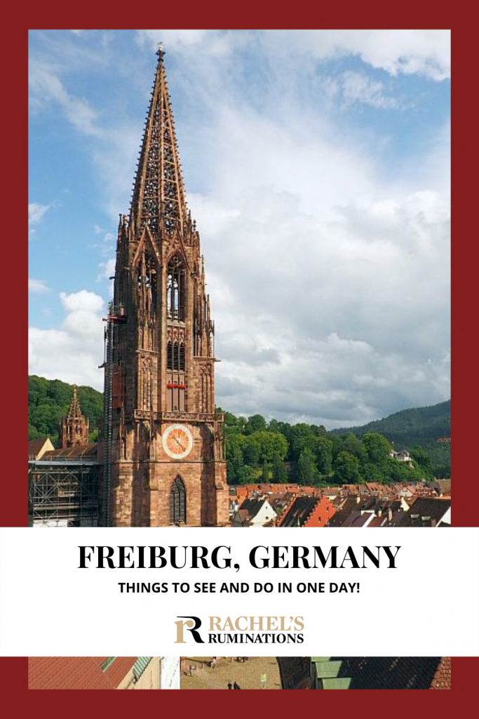 Text: Freiburg, Germany: Things to see and do in one day! (and the Rachel's Ruminations logo). Image: the tower of Freiburg Cathedral, with just the roofs of the town visible to the right and a tree-covered hill behind under a partly cloudy sky.