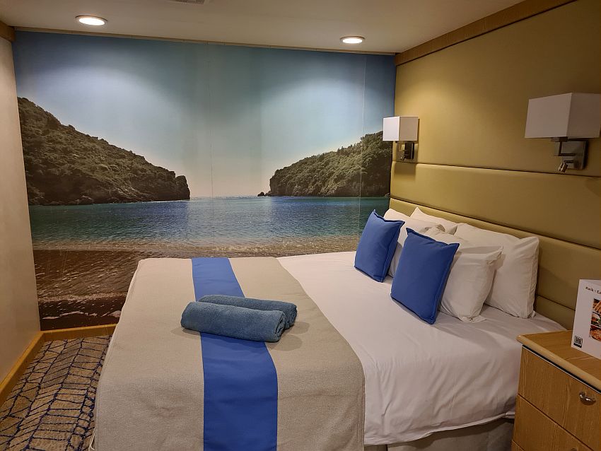 This room on the Celestyal Crystal is square and is filled mostly with a queen-sized bed  extending out from the right-hand wall and a nightstand beside it. The back wall has a photo that covers the entire wall showing a beach in the foreground, a bay, and two hills jutting out from the left and the right to the opening of the bay.