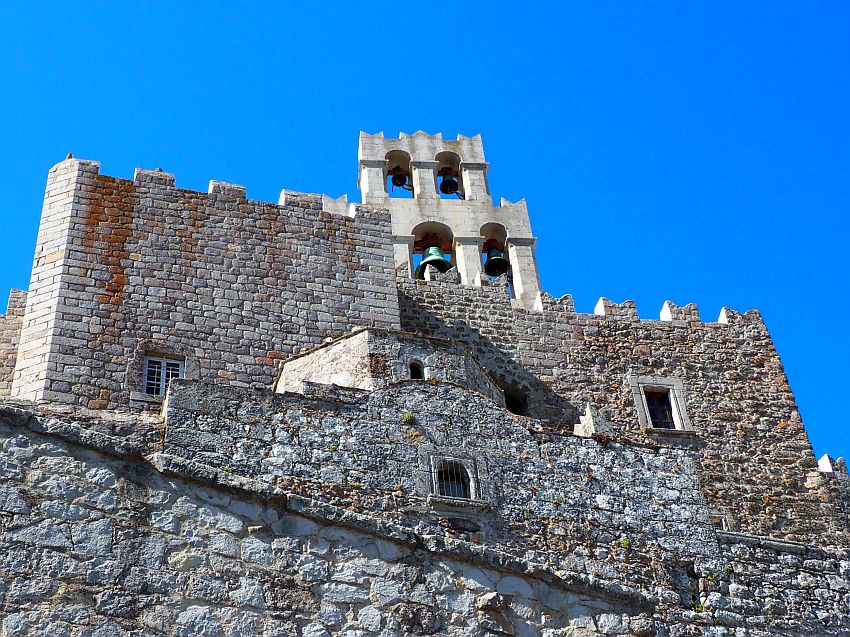 Stone buildings, one in front of the other. The top has crenellations and a bell tower extends up above that, with rounded arches, each with a bell hanging under it.