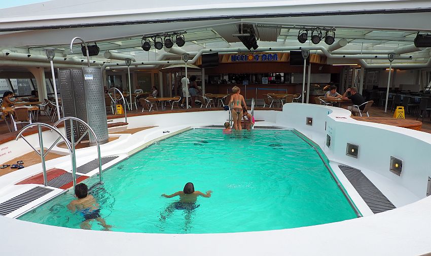 The pool has high walls around it because of sloshing water. The walls are white and the water is bluish-green. Two kids swim in it and a woman at the far end is climbing a ladder into the pool. In the background is a bar, with tables and chairs between it and the pool.