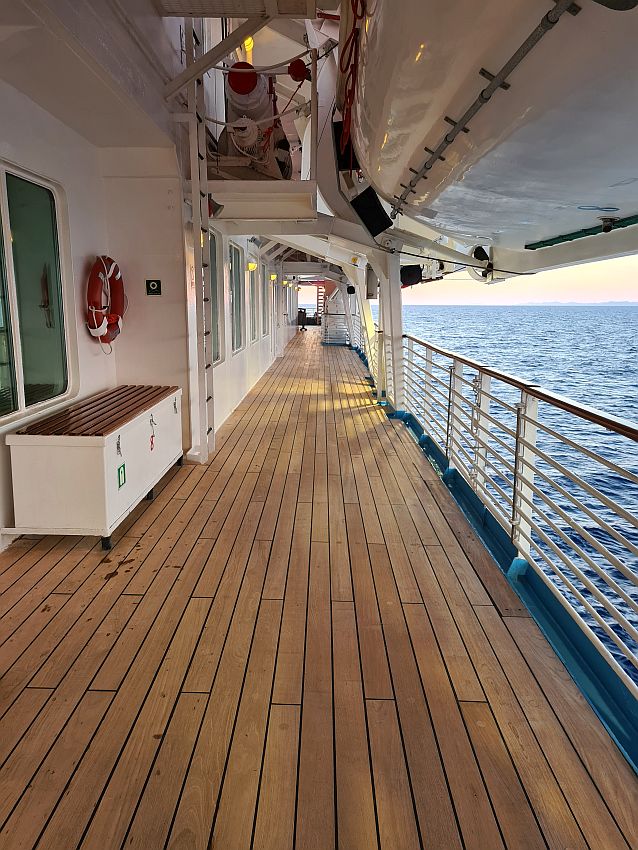 Looking down the long, narrow promenade. Wooden flooring, the rail on the right, the bottoms of lifeboats above.