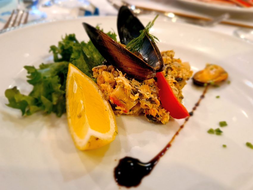 A seafood salad with a mussel shell on top of it. The mussel itself is on the plate beside the seafood salad. A lemon and a sprig of green lettuce beside the salad.