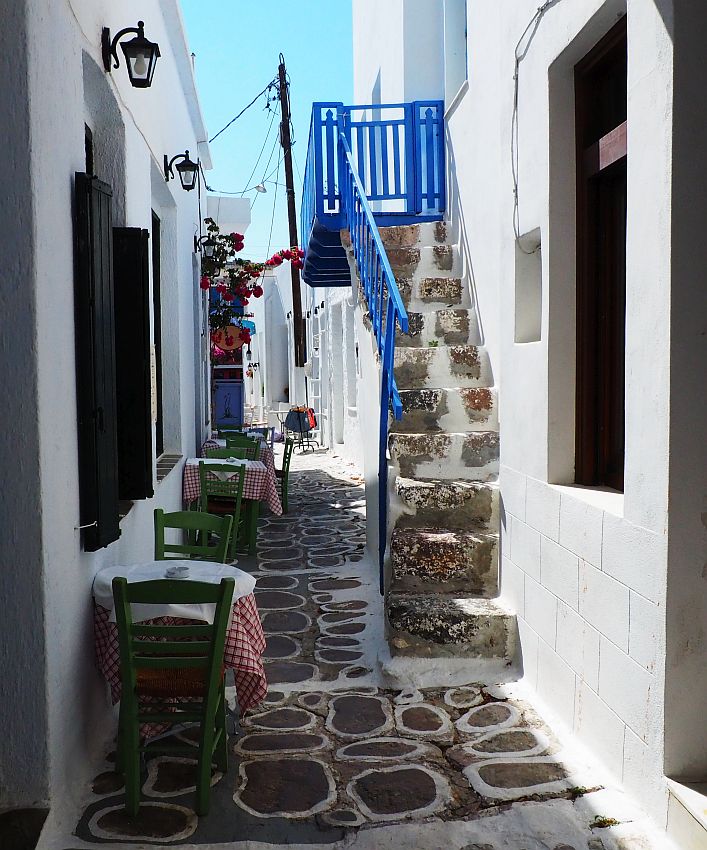 Looking along a very narrow street. On the left, a few small tables against the left wall, each with two chairs against the wall as well. On the right, a stairway up, with the railing painted bright blue, and also the landing at the top of the stairs. All the walls along the street are painted bright white. The ground is flat stones, with white painted around the edge of each. There is just enough space for one person to pass between the tables and the stairway.