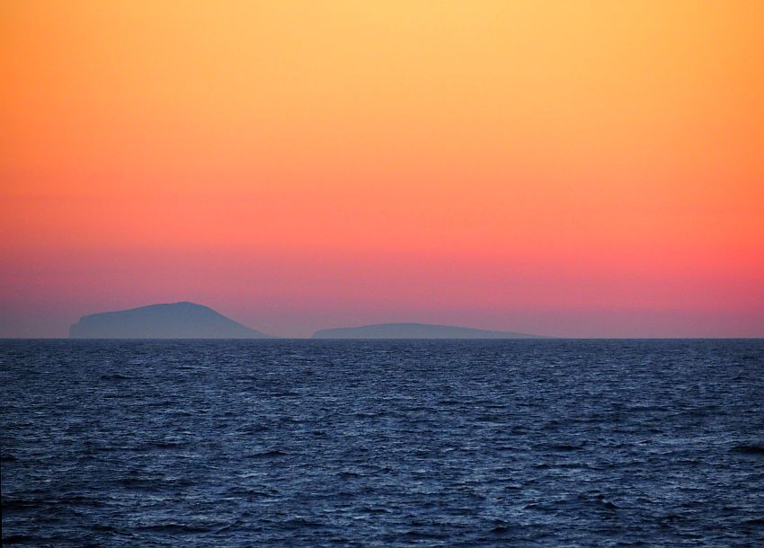 The sky just after the sun is fully down: a few low hills in the distance. Dark sea, then shades of red near the horizon, lightening to orange above.