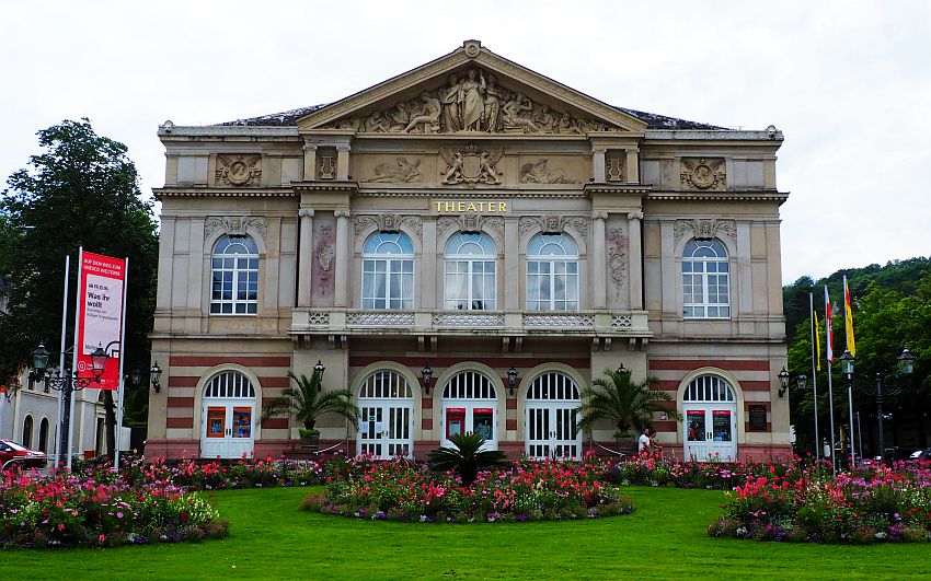 The theater in Baden-Baden, one of the better-known European hot springs towns. It is neo-classical, with pillars between the windows, which have arched tops. The whole building is about 3 stories tall and symmetrical, with 5 windows across on ground and 1st floor. The top floor has an ornate pediment with detailed baroque style statues and bas-reliefs. In front of the building are some neat and colorful flower beds surrounded by grass.