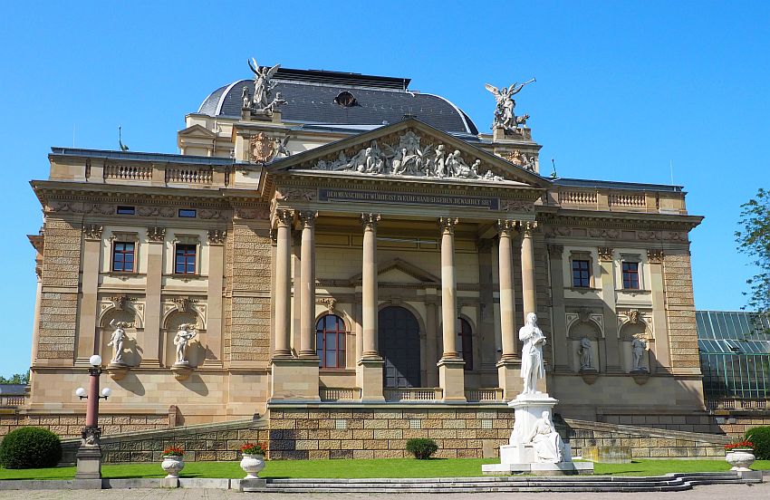 A neo-classical facade, with a center pediment held up by tal pillars. Baroque bas-relief in the pediment, large statues of angels blowing trumpets on each corner above the pediment. Statues in window-sized niches on the lowest story too. In front, a statue of Schiller on a tall pedestal. Typical of the European Thermal Towns.