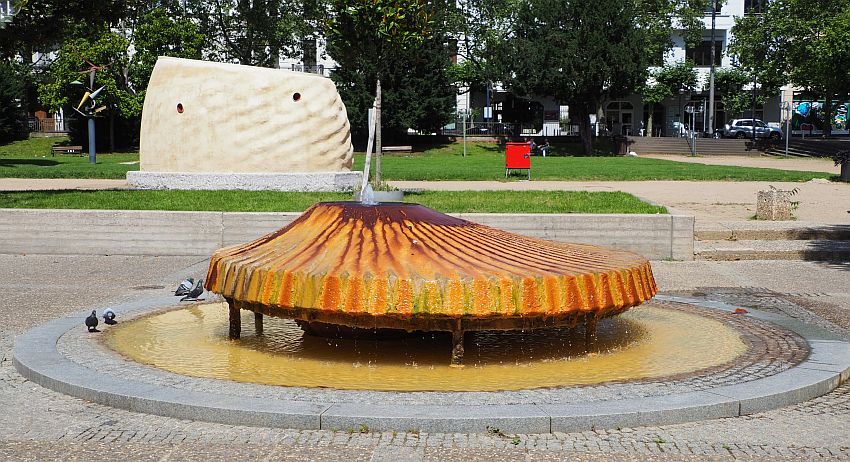 The fountain is round, with the water bubbling out of a hole that is off-center on top, running down the sloping sides on all sides. The slope and the sides where it drips off the fountain into a pool below are striped in yellow and orange. The water in the pool below is yellowish brown.