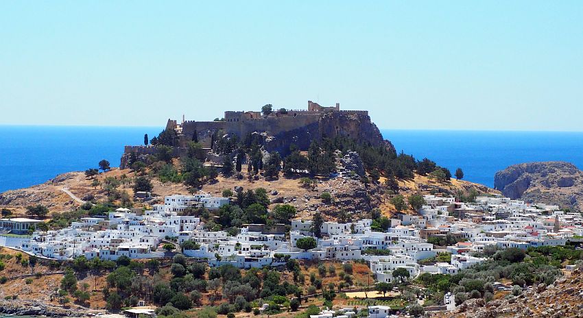 The village of white houses clusters around the base of a hill, which is dark on the sides with pine trees. On top os flat, with stone walls and a few columns visible. Beyond the hill is the blue sea.