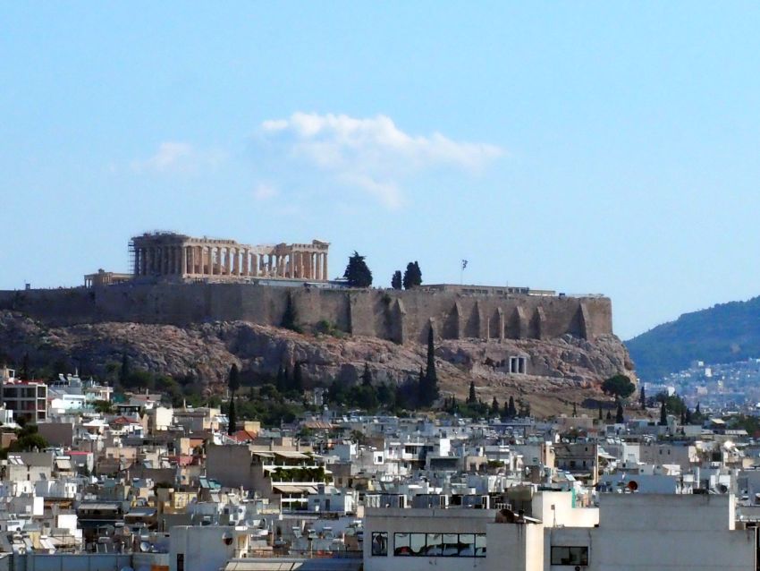 Looking at the Acropolis from a medium height, so that the whole Parthenon can be seen from the side, with it's intact walls of pillars. Below that, the side of the hill is surrounded by stone walls, and below the walls, cliffs. Below the cliffs is the jumble of city buildings.