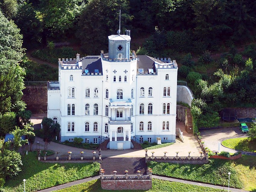 A square block of a building in Bad Ems, Germany, seen from a higher elevation: white, with 4-5 stories, more or less symmetrical. Along the edges of the roof are small crenellations and in the center is a squat round grey tower.