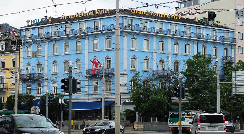 The building is bright blue and stands on a street corner. It is five stories tall, and the top floor has a shallow balcony around it. Here and there some of the lower floors have them as well.  In front, cars pass.