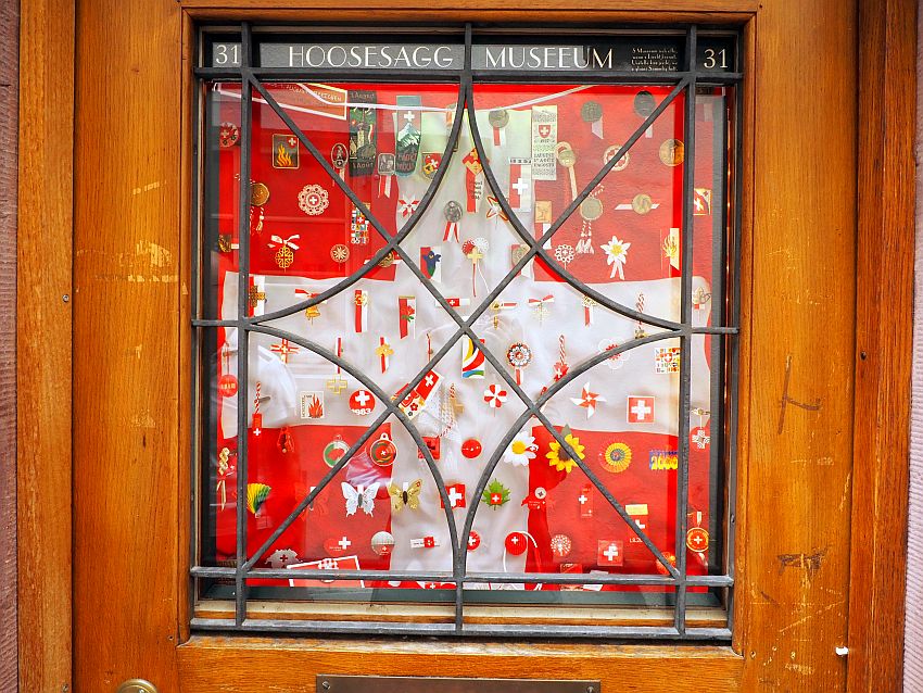 A window says Hoosesagg Museeum at the top and the number 31. A cloth covers the window behind its glass; the cloth shows the swiss flag: red with a big white cross in the middle. On the cloth are many small items: ribbons and flowers and such, almost all in red and white and many showing the same Swiss cross.