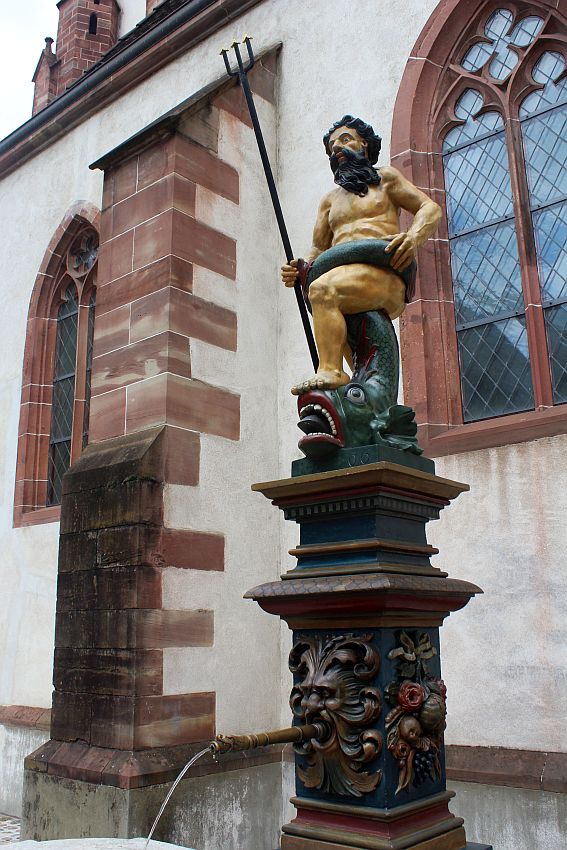 A naked, bearded Poseidon holds a trident. One foot is on top of a snake's head - or perhaps it's a sea serpent or eel. The eel's mouth is open and its tongue is sticking out. It's tail is wrapped between Poseidon's legs and around his waist. Below his statue is the fountain itself: a pipe emerging from a person's mouth.