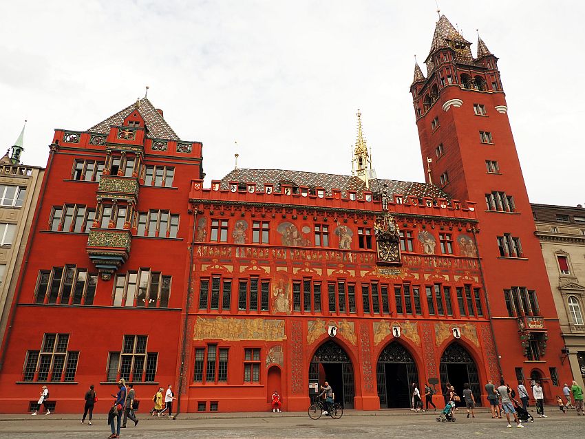 One of the best known of the Basel sights, the Rathaus is painted bright red. It has three gothic arches on the ground floor for its entrance while the rest of the windows are all simple but very narrow rectangles. The left side of the building has the center windows on the upper stories extending outwards like bay windows. The right sid of the building has a tall tower, with fanciful turrets at the top. The building has decorative elements - paintings and sculptures - across the middle, and the roof is colorfully tiled. 