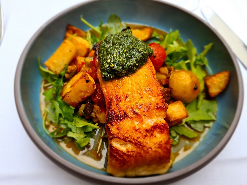 A plate of food. Salmon with a green sauce at one end. The salmon sits on some mixed stewed vegetables and roast potatoes, and it all sits on a bed of rucola or similar.