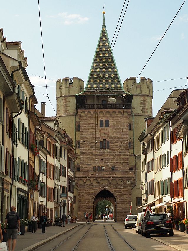 Looking down a street with buildings on either side (white with rows of windows, each with shutters open). At the end of the road, the gate is large, several stories taller than the buildins and blocking the entire road. The tower has an archway at the road level to the other side. The tower is made of stone blocks and has just a few windows on each level. The roof is very steep and pointed and covered in green tile with yellow flowers. On either side of the tower are two more towers, somewhat shorter; they are cylindrical with crenellations at the top.