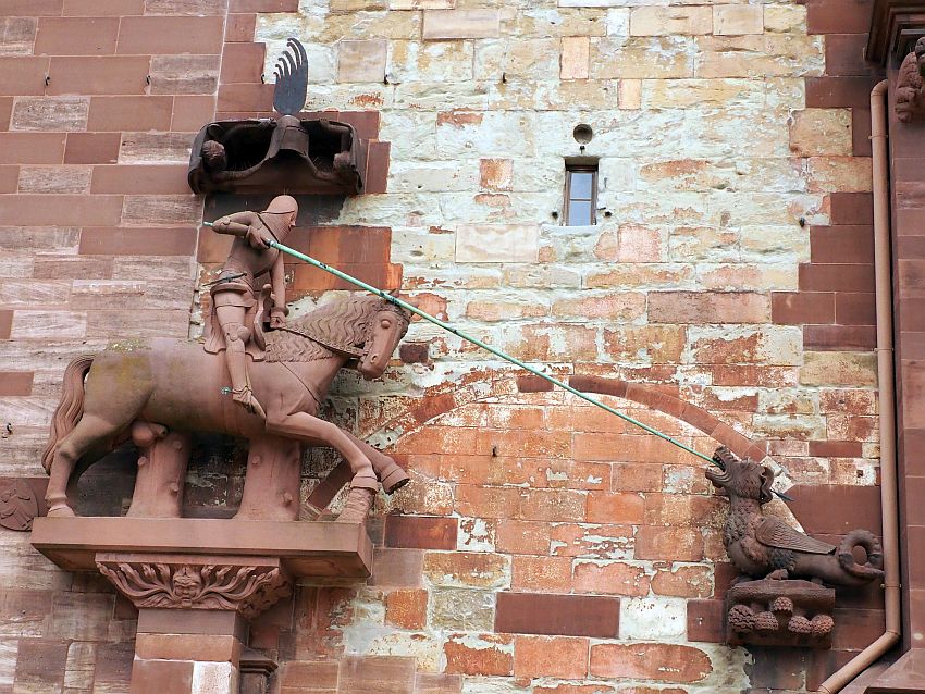 Against a reddish stone wall where the bricks are clearly a range of ages, the St. George statuel. On the left is a knight in armor (St. George) on a horse, carved from red sandstone. He holds a metal lance, longer than the horse's length. It extends to a separate statue of the dragon, facing him, with the lance in its moutn. It is very small compared to the horse, and has a small curled tail.