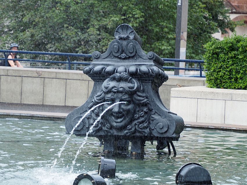 This looks like it might have been a decorative piece on the exterior of a theater. It is triangular, with a face in its middle, with long curly hair. The face is smiling, and water spurts out of its eyes into the pool surrounding it.