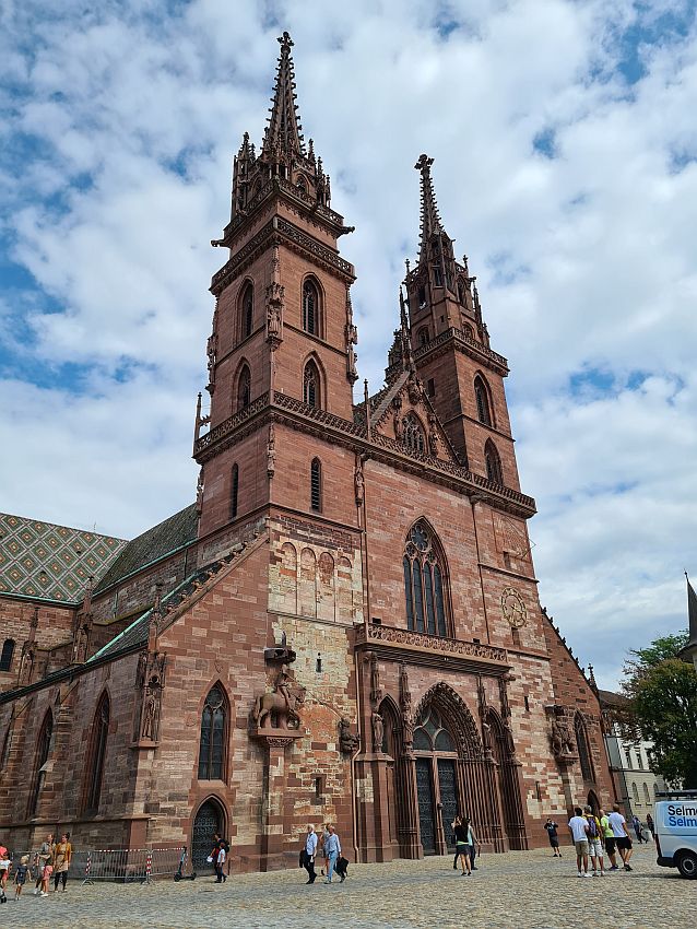 The cathedral's front, seen from an angle. Big entrance right in the middle, and the St. George statue to the left onf the entrance. The entrance and the big window above it have gothic arches. Above that, two spires, not quite matching. Both are square, with a pointed top. 