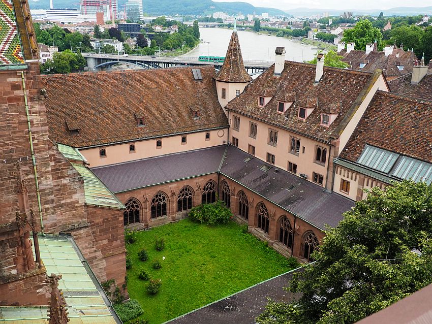 Seen from a couple of stories up, the cloister is square, with green grass and a few bushes in the center. Around the grassy open space is the cloister itself: large openings line the sides looking out on the green. The all have gothic arches and two pillars holding up lacy carved elements in the arch. Behind the cloister are the old monastery buildings, in this picture 2-3 stories higher than the cloister: very plain, light brown, with dark brown roofs and a small turret at the corner.