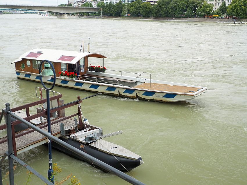 The boat is several meters away from the dock, which is partly visible at the left bottom corner. The boat is long and narrow, and has a cabin at the back 1/3 of the boat with a curved roof over it. The open part in front of the cabin is empty of people.