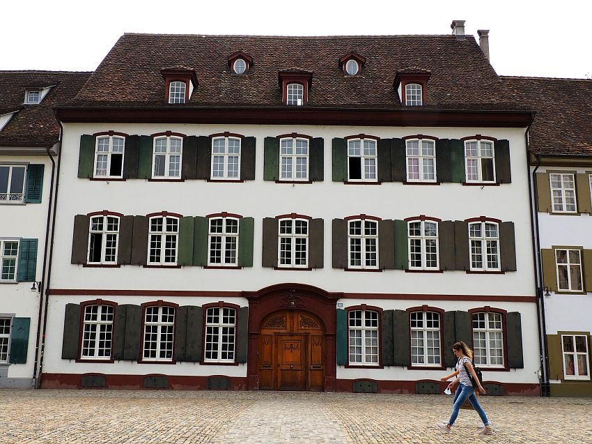 A typical Basel house: simple white plaster, with rows of windows, each with green shutters open around it. The door is large and arched right in the center of the front. Three stories, plus one in the roof. Exactly symmetrical, with 7 windows across each of the upper stories. 